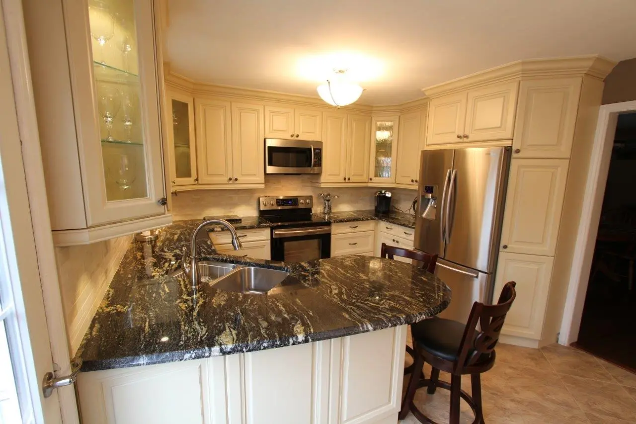 Modern kitchen with cream-colored cabinets, dark marble countertops, stainless steel appliances including a refrigerator and microwave, a built-in stove, a sink with a curved faucet, and two wooden chairs at a peninsula counter.