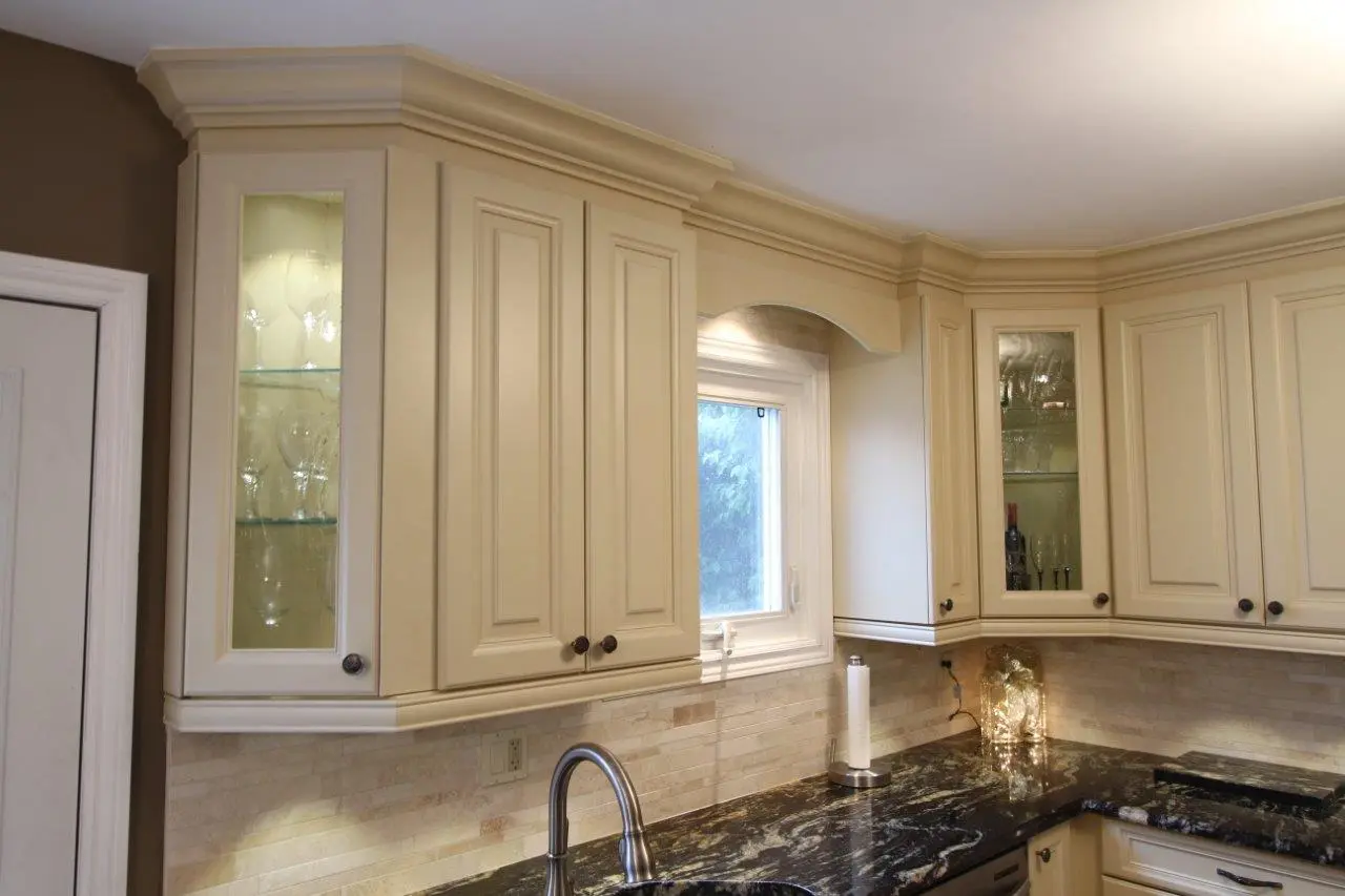 Beige kitchen cabinets with glass-front doors displaying glassware, a small window above a dark granite countertop, a silver kitchen faucet, and a paper towel holder on the counter under soft lighting.