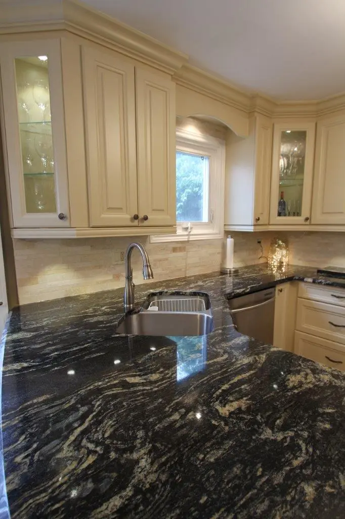 Kitchen with cream-colored cabinets and dark granite countertops featuring beige and white swirls, a stainless steel sink with a modern faucet, a dishwasher, and under-cabinet lighting, with a window above the sink.