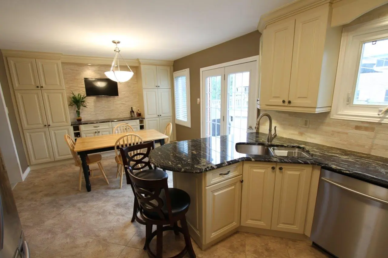 Spacious kitchen with beige cabinets, black granite countertops, a corner sink, stainless steel dishwasher, a wooden dining table with chairs, and a mounted television on a beige tiled backsplash wall, with glass doors leading outside.
