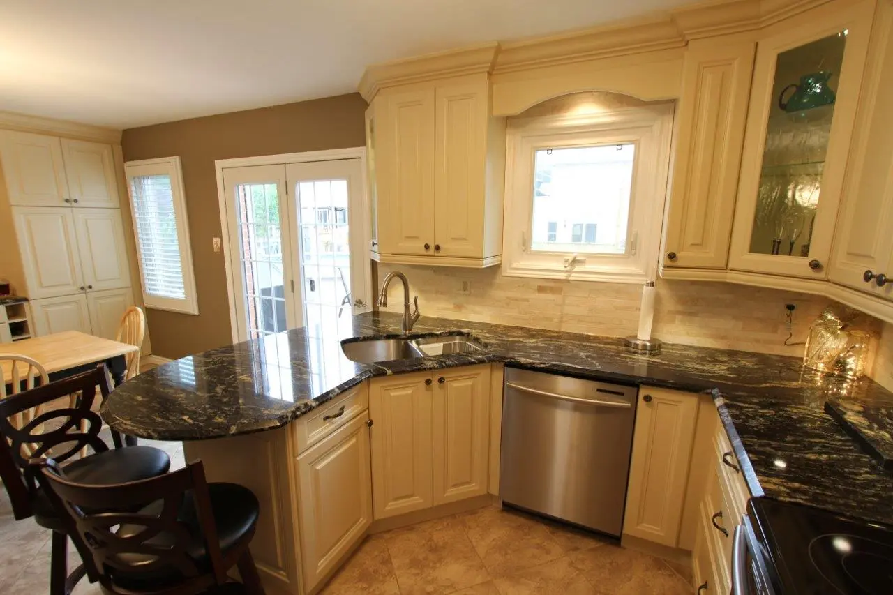Bright kitchen with cream-colored cabinets, black granite countertops, a double sink under a window, a stainless steel dishwasher, dark wooden bar stools, and a dining table near glass-paned double doors leading outside.