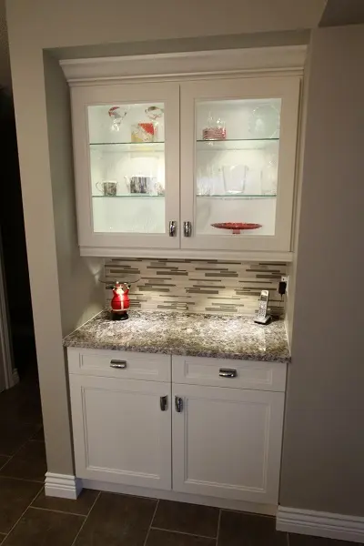 Built-in white kitchen cabinet with glass doors displaying glassware, a granite countertop with a small red and white candle holder, tiled backsplash, and a cordless phone on the right side under soft lighting.