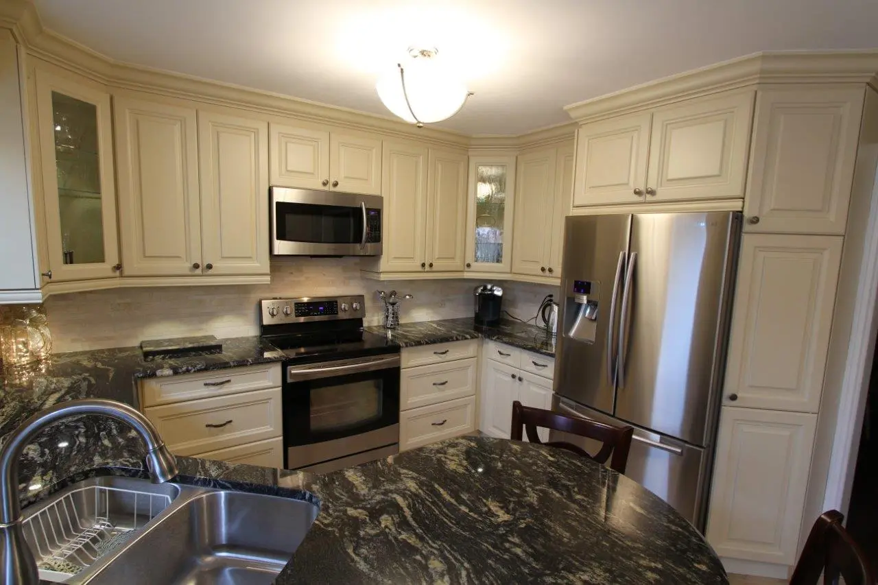 Modern kitchen corner with cream-colored cabinets, black and gray granite countertops, stainless steel sink, stove, microwave, and refrigerator, and dark wood chair partially visible.