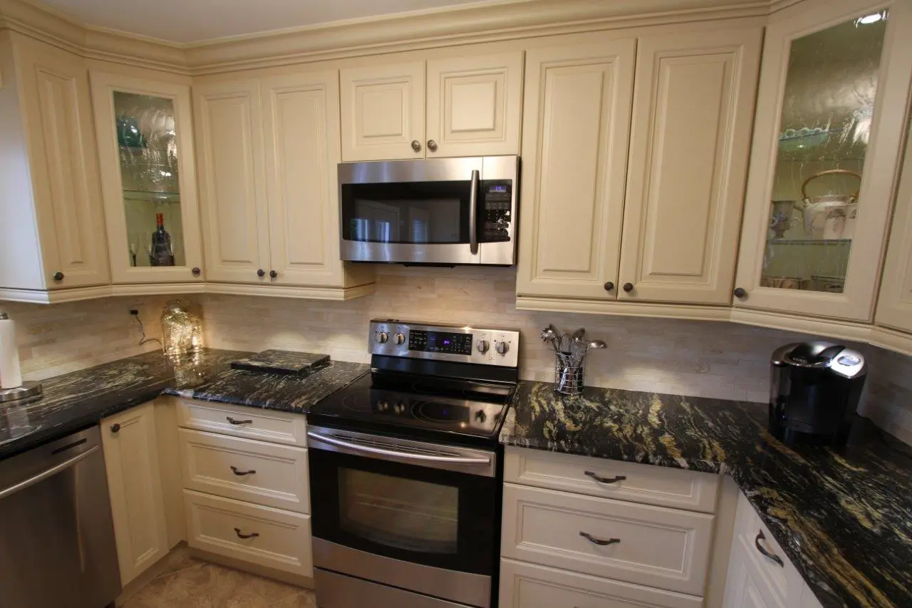 Modern kitchen corner with stainless steel electric stove and microwave, cream-colored cabinets with glass inserts, and dark granite countertops with gold and green patterns, featuring a coffee maker and utensil holder.