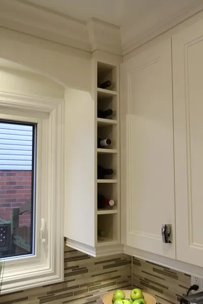 Narrow vertical wine rack built into the corner of cream-colored kitchen cabinets, holding several wine bottles, next to a window with a view of a brick wall and a wooden fence, above a countertop with a bowl of green apples and a mosaic tile backsplash.