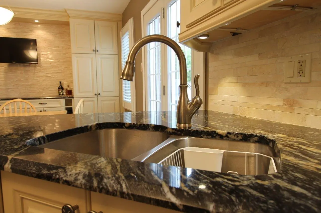 Close-up of a kitchen sink with a curved brass faucet mounted on a dark granite countertop, beige tiled backsplash, cream-colored cabinets, and a dining area with wooden chairs and a wall-mounted TV in the background.