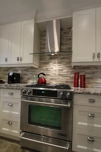 Modern kitchen with a stainless steel Jenn-Air stove and oven, a red kettle on the stove, white cabinets with silver handles, a patterned tile backsplash in neutral tones, and a coffee machine and red canisters on the granite countertop.