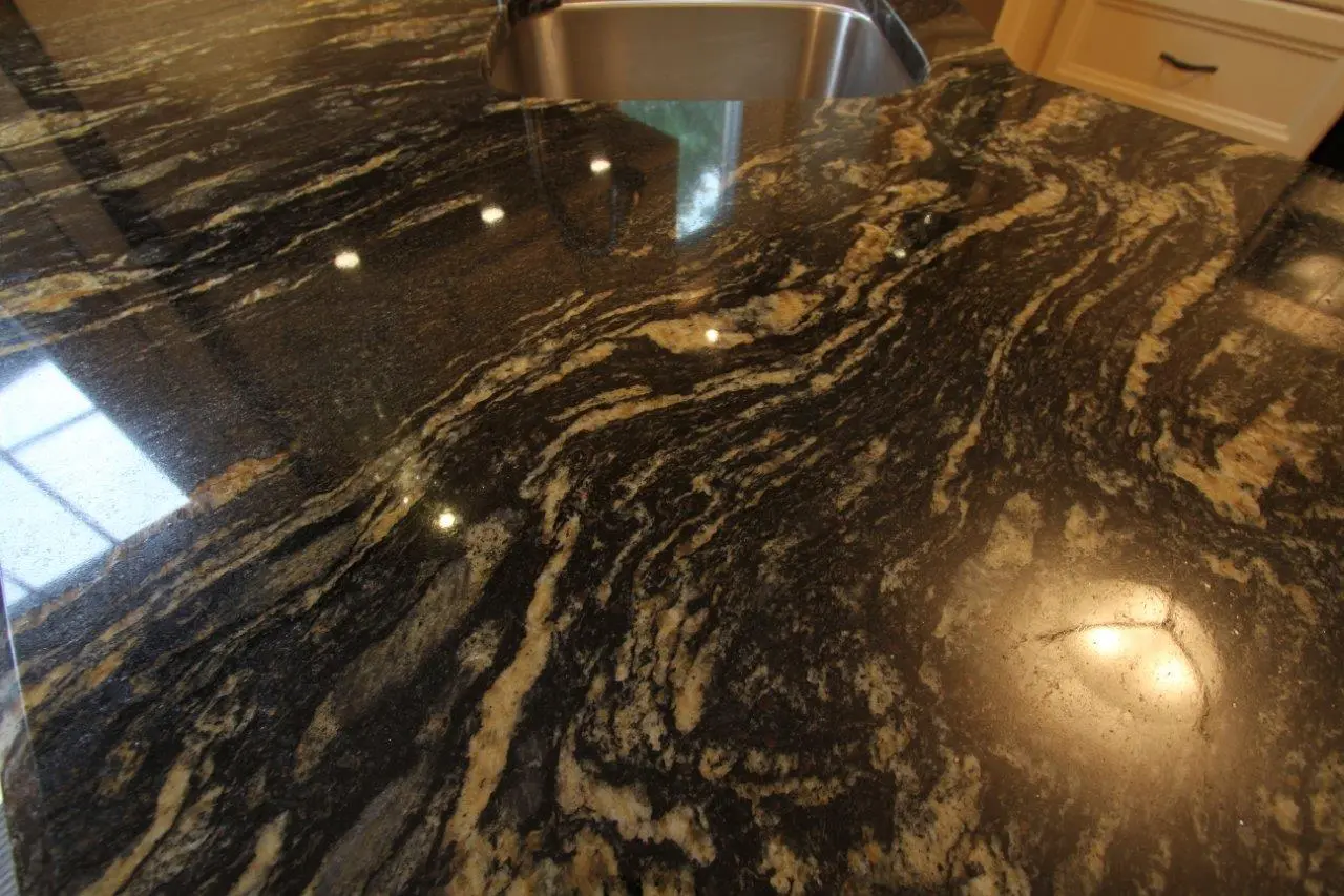 Close-up of a polished granite kitchen countertop with black, gray, and beige swirling patterns beneath a stainless steel sink and reflections of ceiling lights.