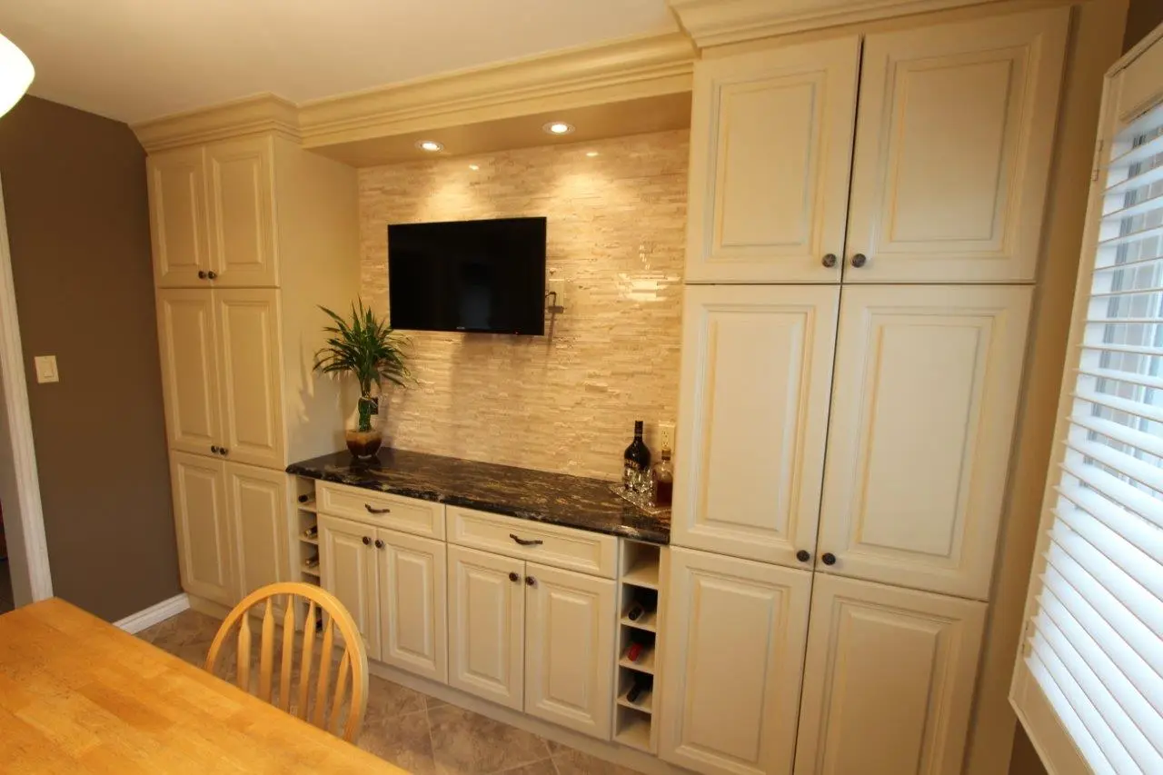 Kitchen area with cream-colored cabinets, a black marble countertop, a mounted flat-screen TV on a beige tiled backsplash, a potted plant on the countertop, a bottle of liquor with glasses, and a wooden dining table with a chair partially visible in the foreground.