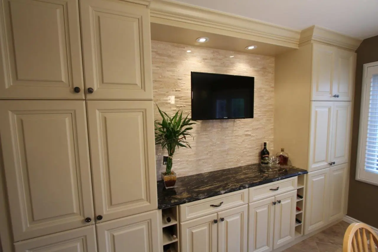 A modern kitchen or bar area with cream-colored cabinets, a black marble countertop, a mounted flat-screen TV, a beige textured stone backsplash, a potted plant, and bottles of liquor on the counter, illuminated by recessed ceiling lights.
