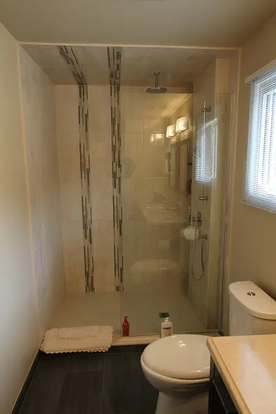 Modern bathroom with a walk-in glass shower featuring beige tiles with vertical decorative strips, a rain showerhead, a handheld shower fixture, a white toilet, a beige countertop, a small white rug, and two small bottles on the shower floor, illuminated by natural light from a window with blinds on the right side.
