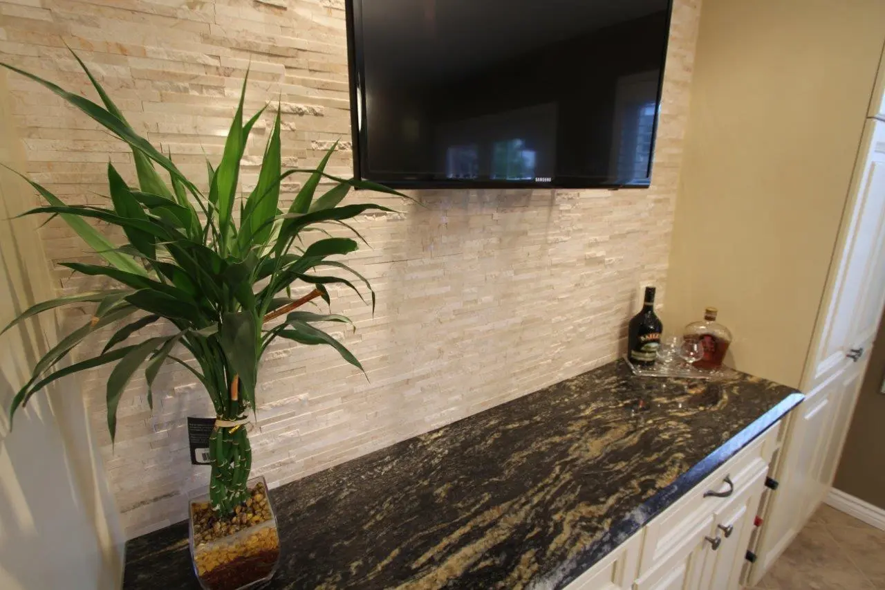 Indoor countertop with a black and gold granite surface, featuring a green potted plant with wide leaves on the left and a small whiskey and liqueur setup with glasses on the right, beneath a mounted flat-screen TV against a beige stone tile wall.