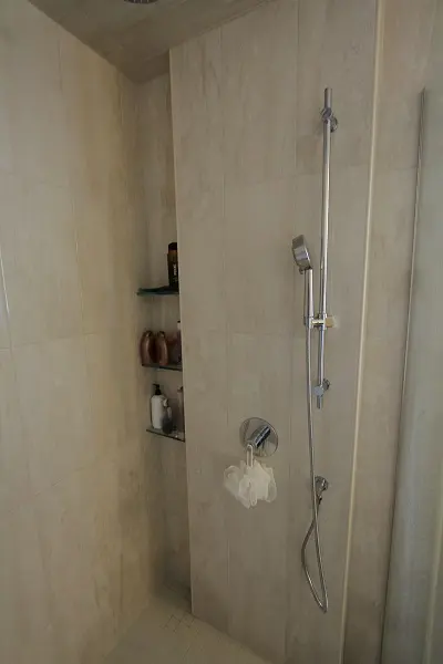 Corner of a tiled shower area with a mounted adjustable handheld showerhead, a chrome control knob, a white bath sponge hanging below the control, and three glass shelves holding various bottles of shower products.