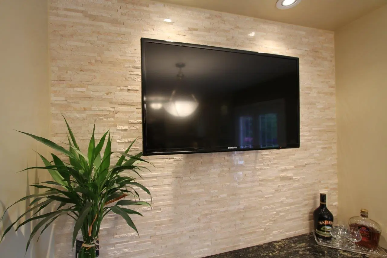 Flat-screen Samsung TV mounted on a beige textured stone wall above a dark granite countertop, with a green leafy plant to the left and bottles of liquor with glasses on the right side of the countertop.