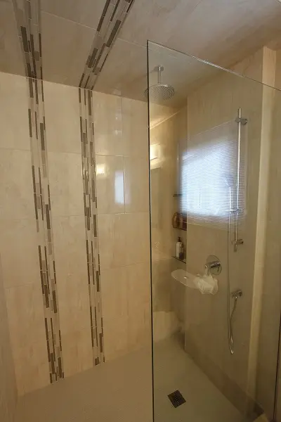 Modern bathroom shower with beige tiled walls featuring vertical mosaic accents, a glass partition, rainfall showerhead, handheld shower attachment, and built-in niche holding toiletries, illuminated by a window with blinds.