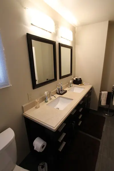 Modern bathroom with a double sink vanity featuring two rectangular mirrors, countertop accessories, and overhead lighting, with dark cabinetry and a toilet partially visible on the left.