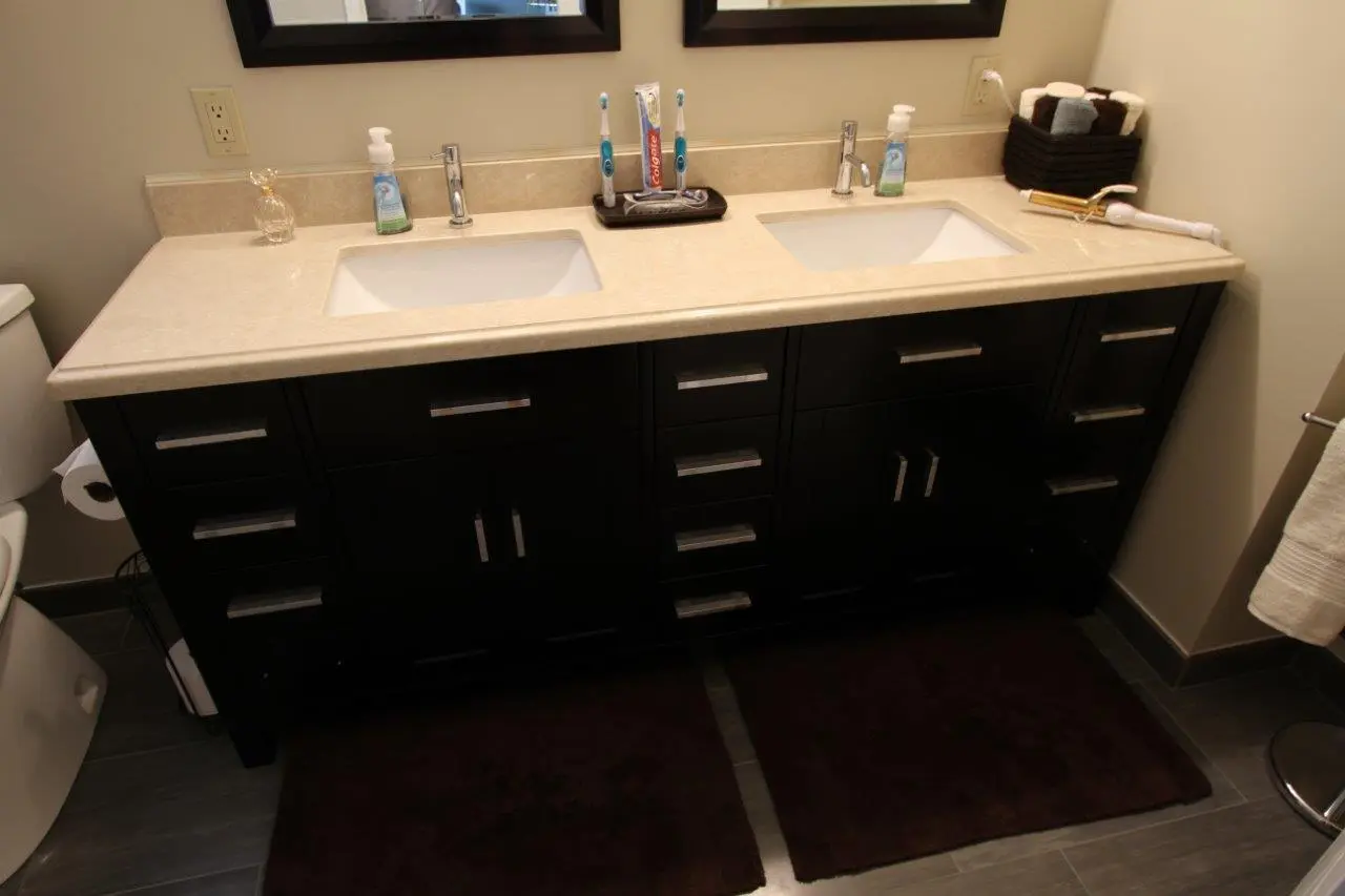 Double bathroom sink with beige countertop and black cabinetry, featuring two toothbrushes, toothpaste, soap dispensers, a basket with rolled towels, and a hair curling iron on the right side, two dark brown mats on the floor, and part of a toilet and towel rack visible.