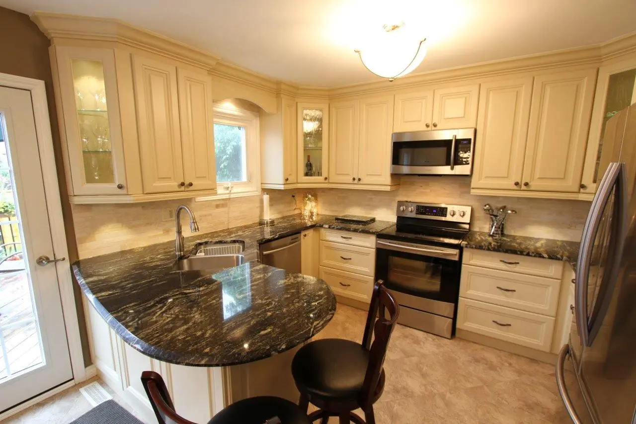 Modern kitchen with cream-colored cabinets, black granite countertops, stainless steel appliances including a microwave, oven, dishwasher, and refrigerator, and two dark wood bar stools at the counter near a glass door leading outside.
