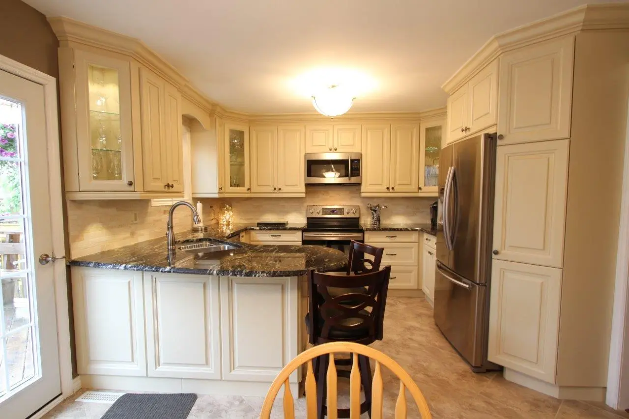 Well-lit kitchen with cream-colored cabinetry, black granite countertops, stainless steel appliances including a refrigerator and stove, a curved island with a sink and two dark wooden chairs, and a glass-paneled door on the left side.