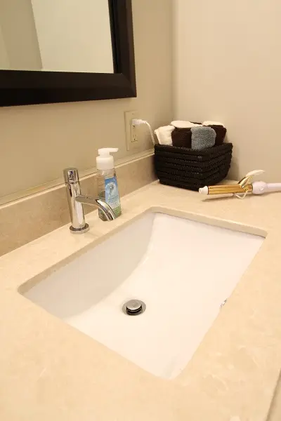 Bathroom sink with a chrome faucet, a transparent soap dispenser, a black basket containing neatly folded towels, and a gold-colored hair curling iron on a beige countertop.