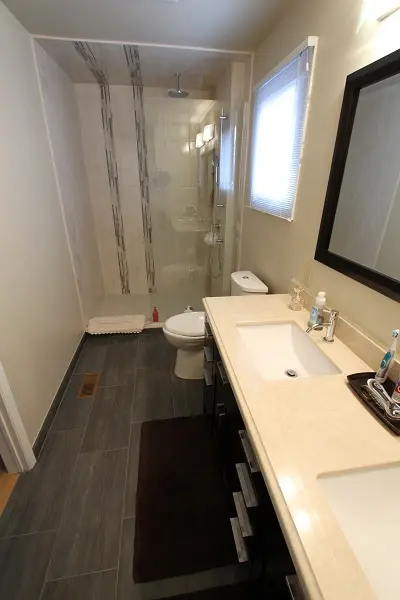 Modern bathroom with beige countertop and double sink vanity, dark wood cabinets, glass-enclosed shower with vertical decorative tile strips, white toilet, window with blinds, large mirror, and dark floor tiles with a brown bath mat.