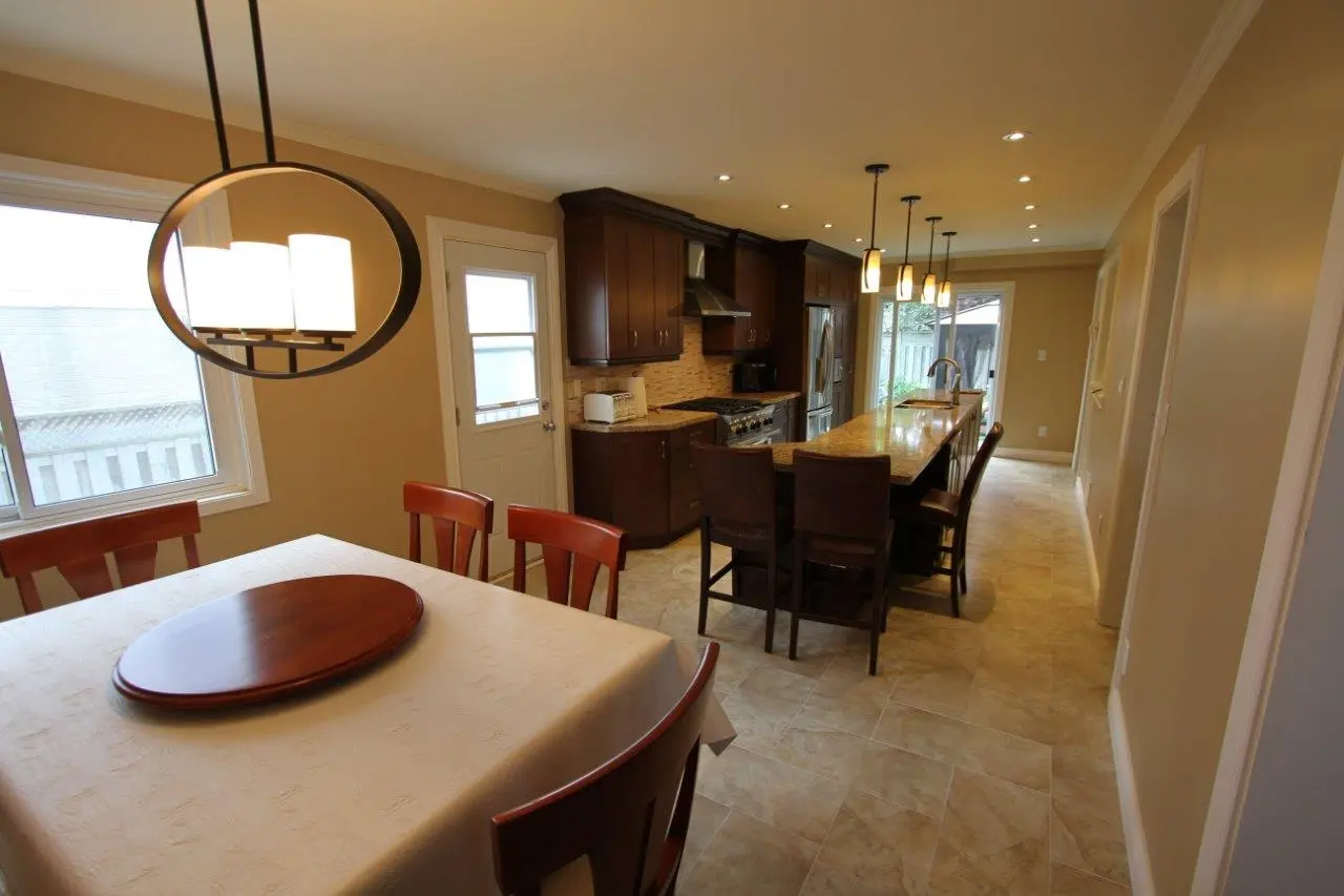 View of a modern kitchen with beige tiled floor, dark wooden cabinets, and a long island with four dark wooden bar stools, pendant lights above the island, and a dining table with a white tablecloth and wooden chairs in the foreground near a window and door.