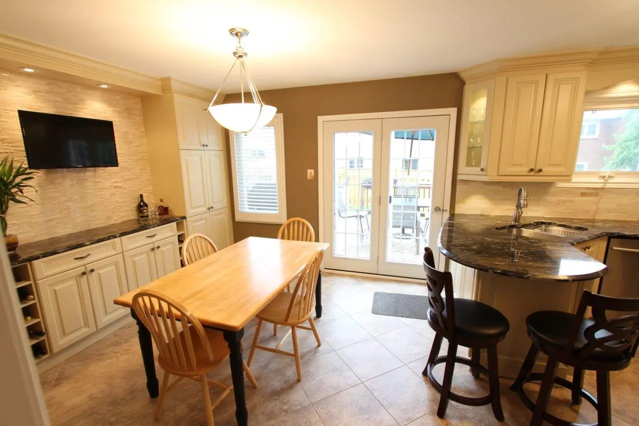 Bright kitchen and dining area with light wooden table and chairs, black granite countertops, beige cabinets, a wall-mounted TV, a large pendant light, and glass doors leading to an outdoor space.