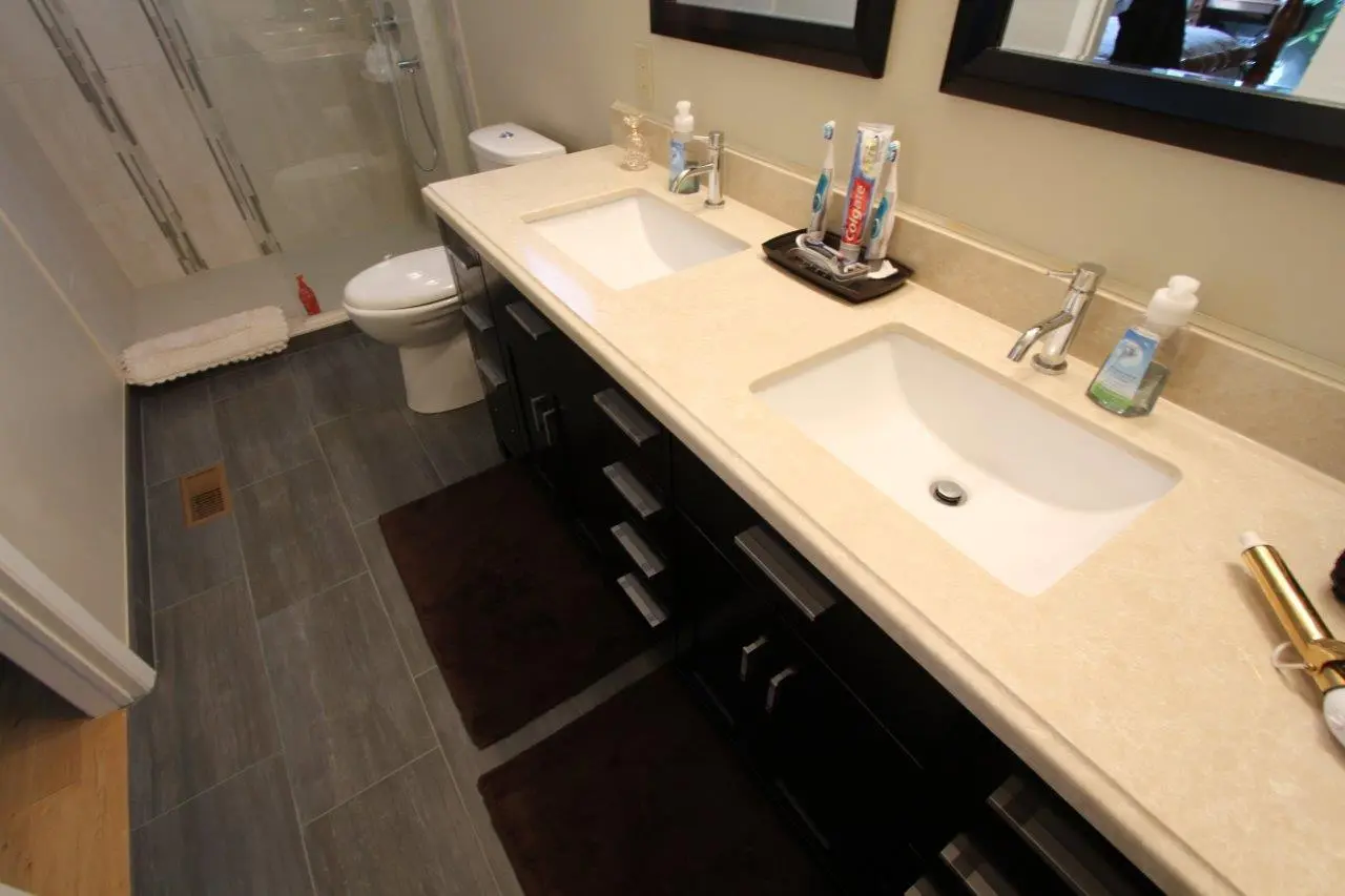 Modern bathroom with dual sinks set in a beige countertop, dark cabinetry, a toilet, a glass-enclosed shower area, and dark brown floor mats on gray tile flooring.