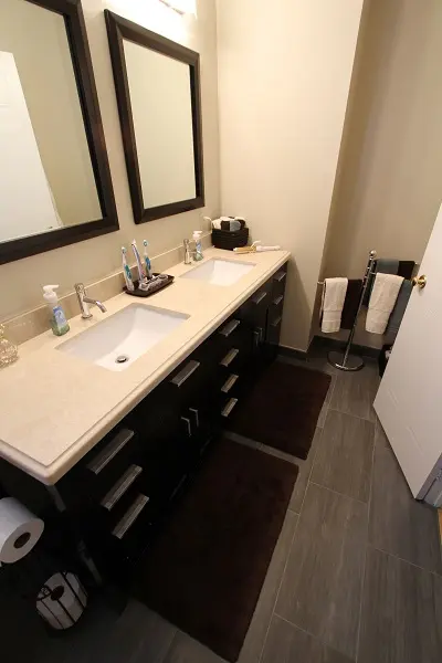 Modern bathroom with a double sink vanity featuring beige countertops, dark wood cabinets, two mirrors, toothbrushes and soap dispensers on the counter, dark brown floor mats, and a towel rack with white towels near a white door.