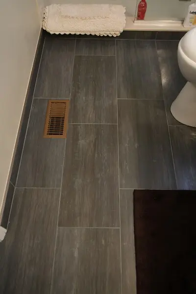 Bathroom floor with large dark gray rectangular tiles, a brown floor vent on the left, a dark brown bath mat on the right, and a section of a white toilet and bath towels in the upper corner.