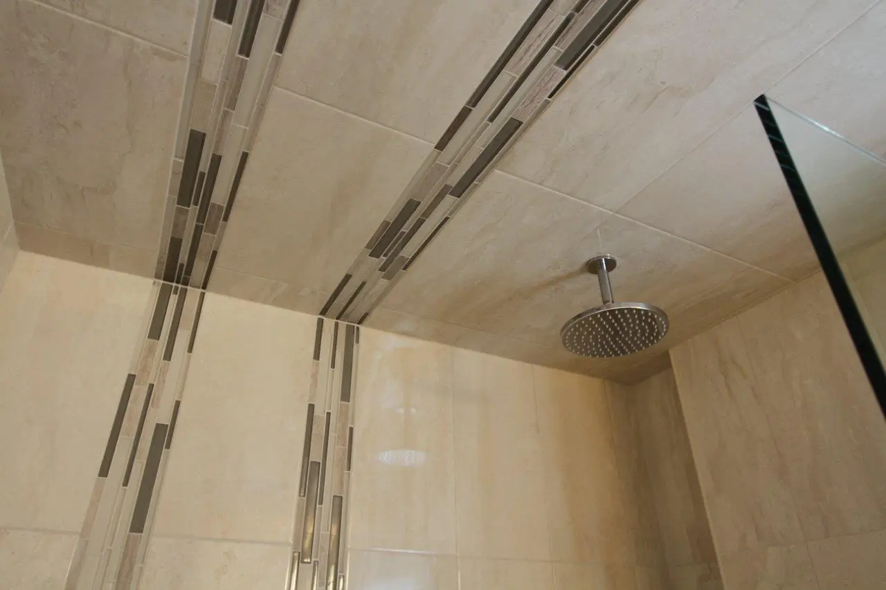 Ceiling view of a shower with beige tiles and vertical decorative strip tiles, featuring a round stainless steel rain shower head.