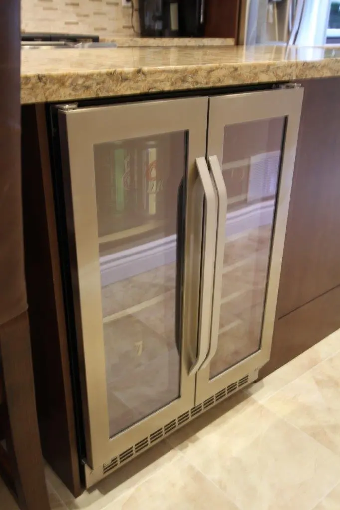 Double-door under-counter beverage fridge with glass panels and metal handles built into a kitchen cabinet with a marble countertop.