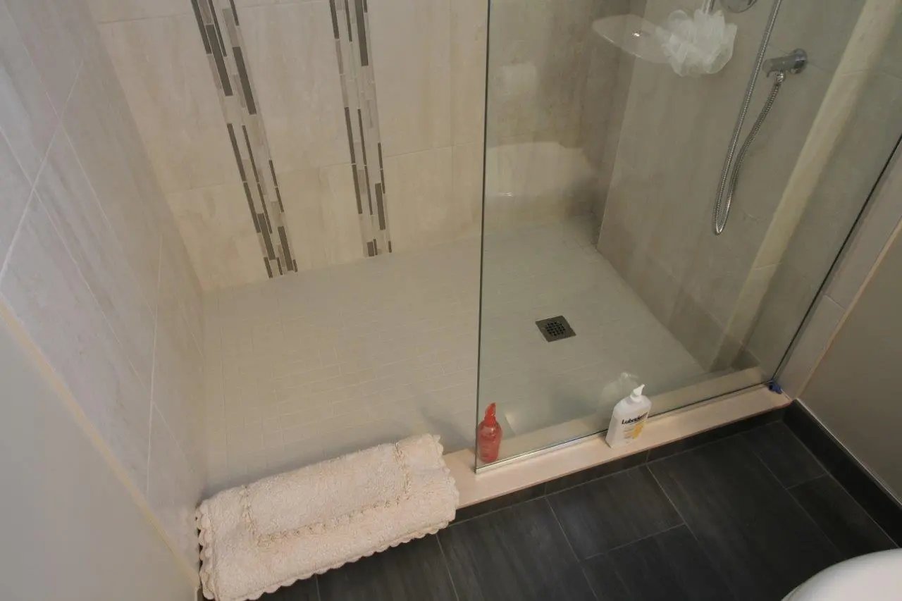 Modern shower area with beige tiled walls and floor, a glass shower door, a chrome handheld showerhead, a red bottle and a white white bottle of Lubriderm lotion placed on the shower base, and a beige bath mat on the dark gray tiled bathroom floor outside the shower.