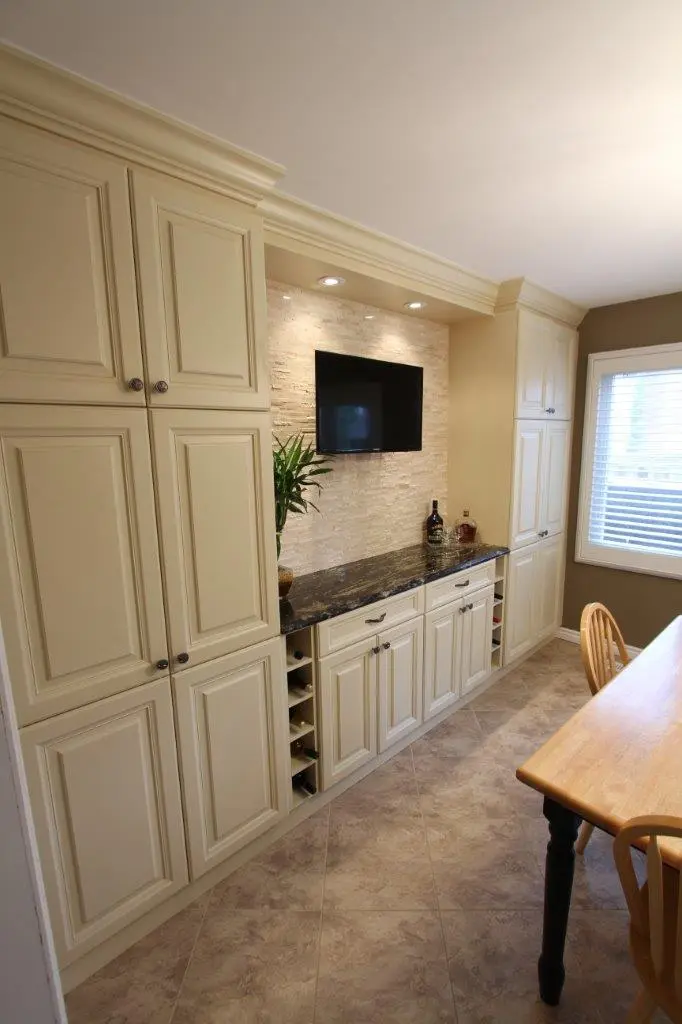 A kitchen or dining area with cream-colored cabinets, a black granite countertop, a small wall-mounted TV, decorative bottles, a plant, and a wooden dining table with chairs, illuminated by recessed ceiling lights.