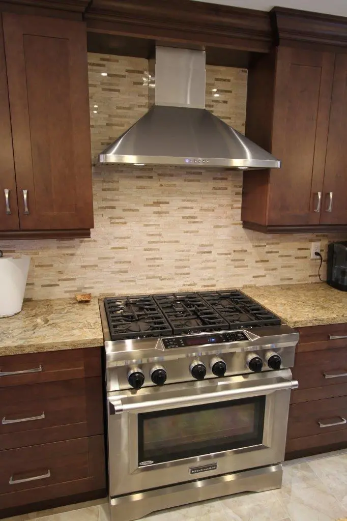 Modern kitchen with a stainless steel gas stove and oven, brown wooden cabinets, beige tiled backsplash, and a stainless steel range hood above the stove.