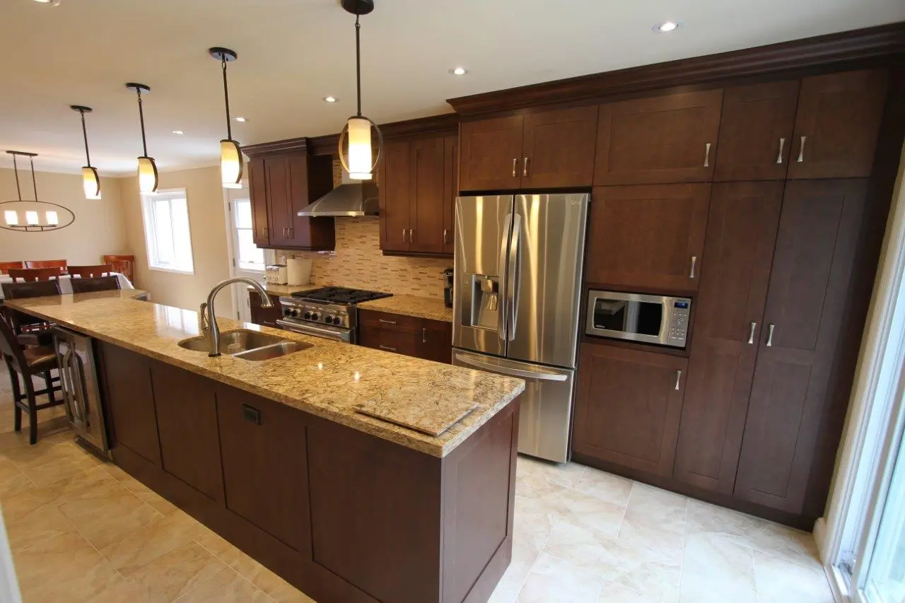 Modern kitchen with dark wooden cabinets, stainless steel refrigerator and stove, granite countertops on a large island with a built-in sink and hanging pendant lights above, beige tiled floor, and a dining area with chairs in the background.