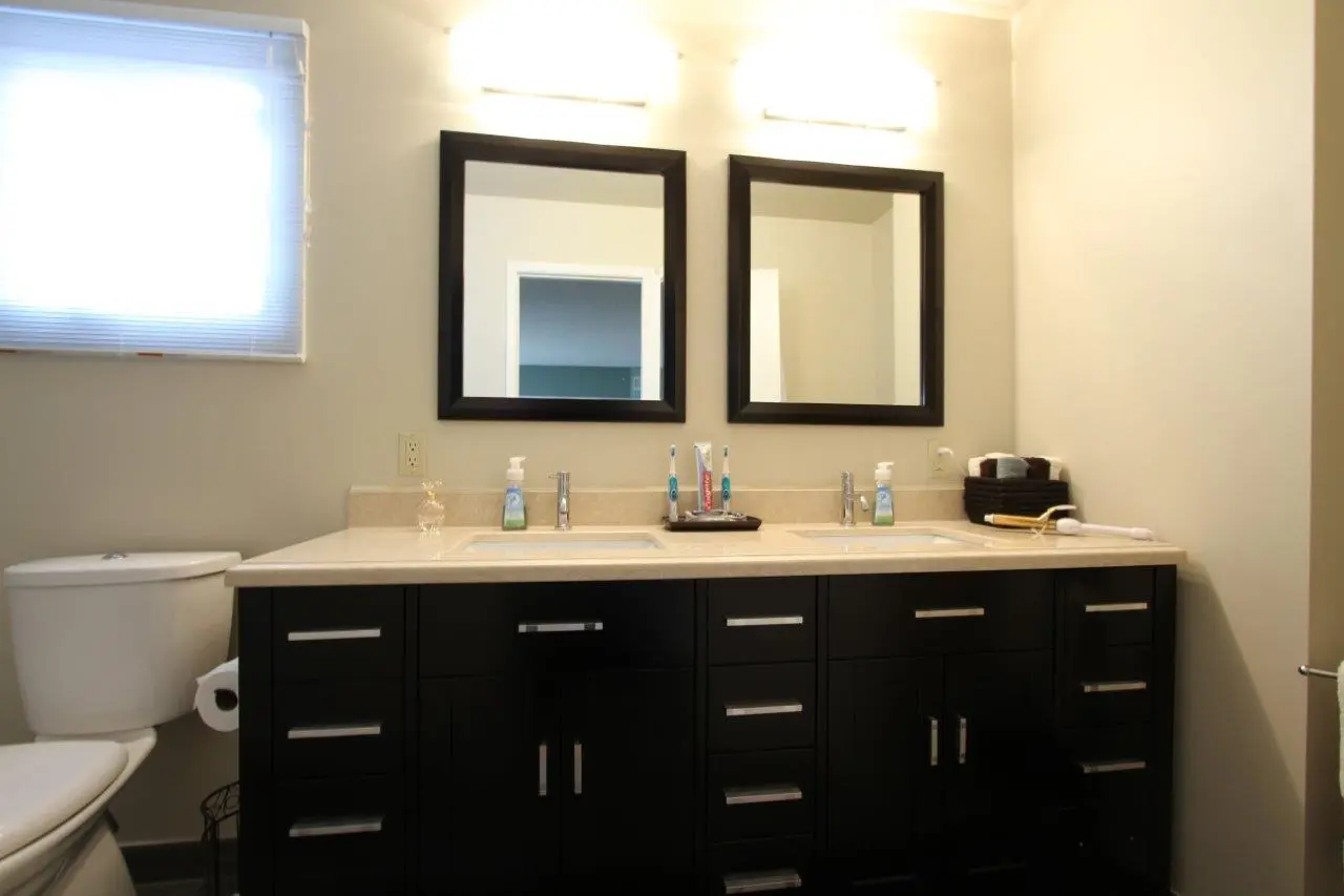 Modern bathroom with a double sink vanity featuring black cabinets and light-colored countertop, two square mirrors with black frames above sinks, a window with blinds on the left, a toilet next to the vanity, and various toiletries such as toothbrushes, soap dispensers, and towels on the countertop.