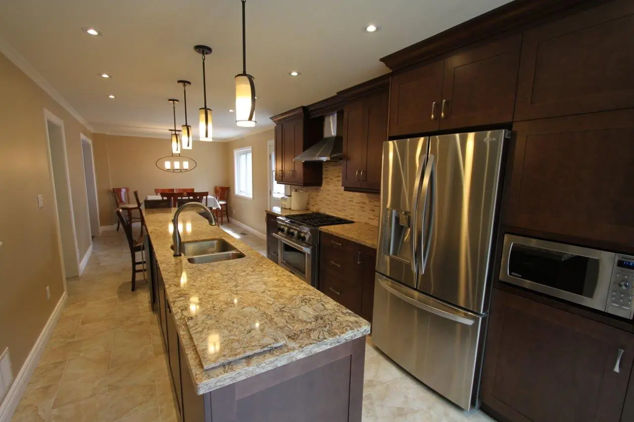Modern kitchen with dark wood cabinets, granite countertop island with double sink, stainless steel refrigerator and oven, pendant lights above the island, and dining table with chairs in the background.