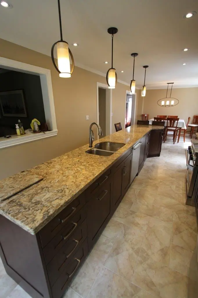 Long kitchen island with granite countertop and dark wooden cabinets, stainless steel double sink, modern pendant lights above, beige tiled floor, and dining area with table and chairs in the background.