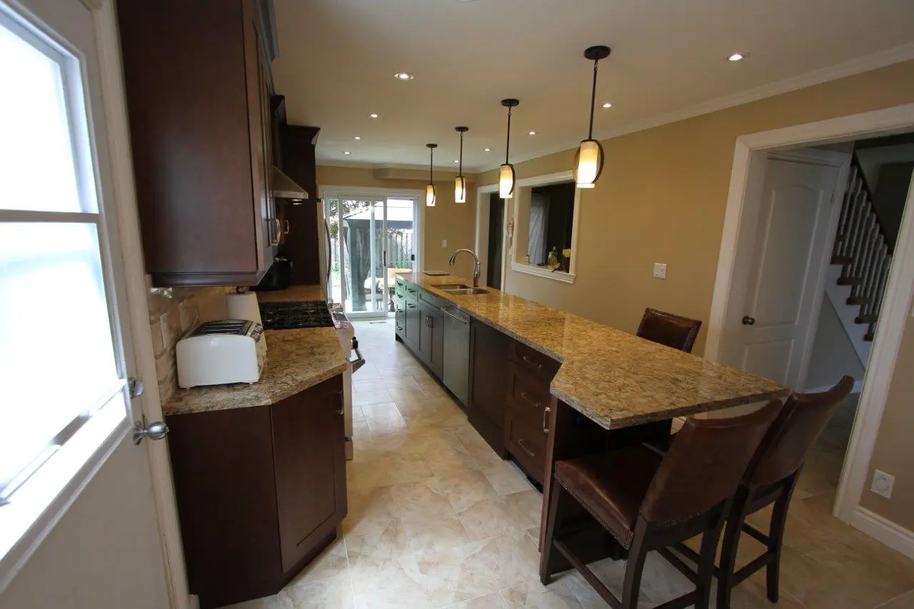 Modern kitchen with beige tiled floor, granite countertops, dark wood cabinets, and pendant lights above a long island with two bar stools, with a door and glass sliding doors at the far end.