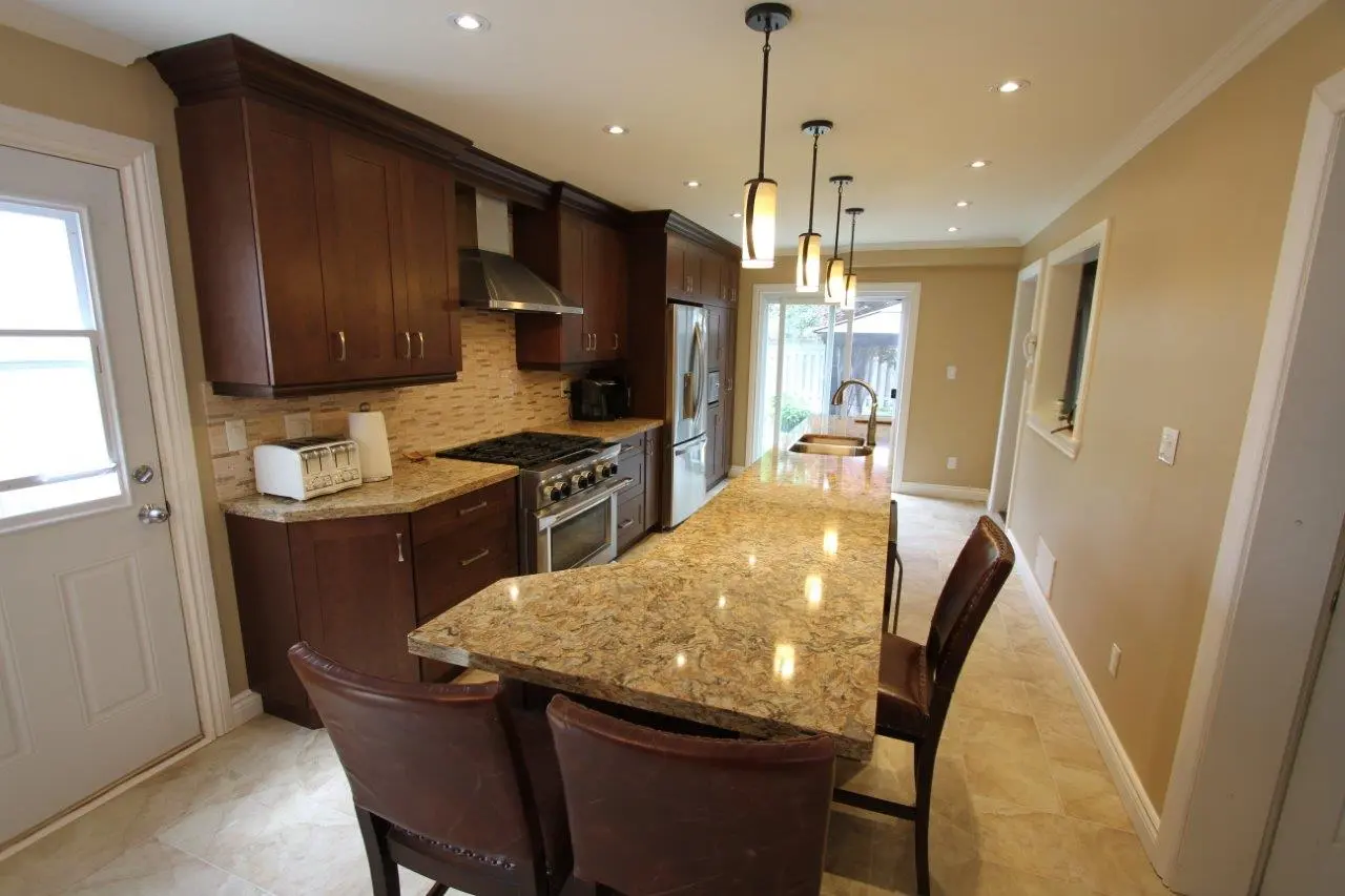 Modern kitchen with granite countertops, dark wood cabinetry, stainless steel appliances, three pendant lights above the island, and three brown leather chairs on one side of the island.