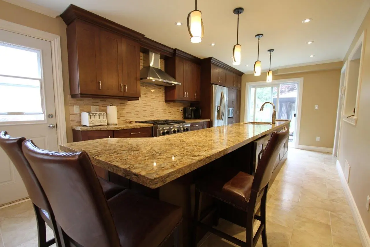 Modern kitchen with a long granite island countertop, dark wood cabinets, stainless steel appliances, three pendant lights hanging from the ceiling, and three brown leather bar stools along the island.