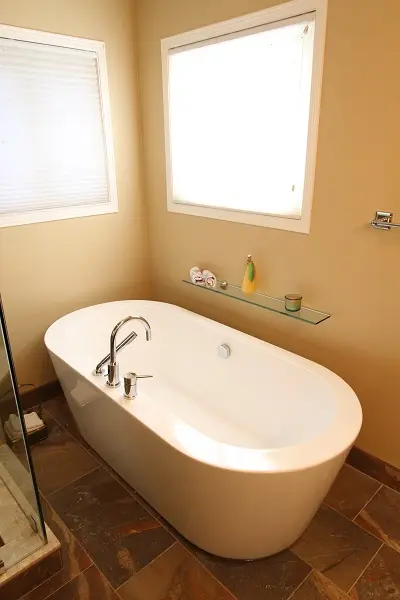 Modern white freestanding bathtub with chrome faucet in a corner bathroom with beige walls, a glass shelf holding small decorative items, two windows with blinds, and brown tiled floor.
