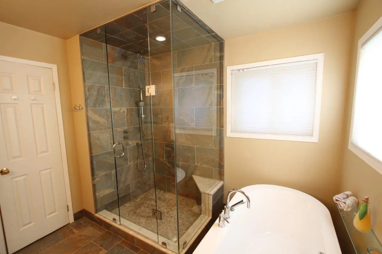 Modern bathroom with a glass-enclosed tiled shower featuring a built-in corner bench and a white bathtub with chrome faucets next to a window with blinds.