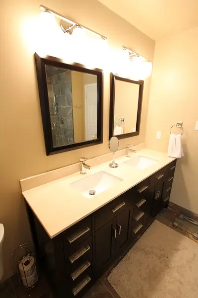 Double sink bathroom vanity with beige countertop, dark wooden cabinets, two rectangular mirrors with dark frames, and three bright lights above each mirror in a tan-colored bathroom