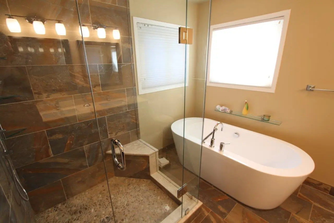 Modern bathroom with a glass-enclosed shower featuring brown stone tiles, a built-in corner bench, and a white freestanding bathtub next to a window with a small glass shelf holding toiletries.