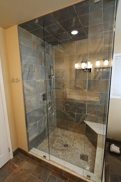 Modern bathroom shower with glass enclosure, stone tile walls and ceiling, pebble floor, built-in bench, and overhead rain showerhead under recessed light