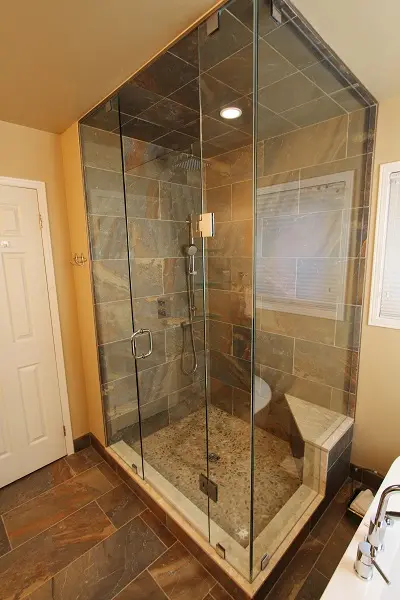 Modern bathroom shower enclosure with glass walls, stone tiled interior, pebble stone floor, built-in corner bench, and chrome shower fixtures, adjacent to a white bathtub and beige walls.