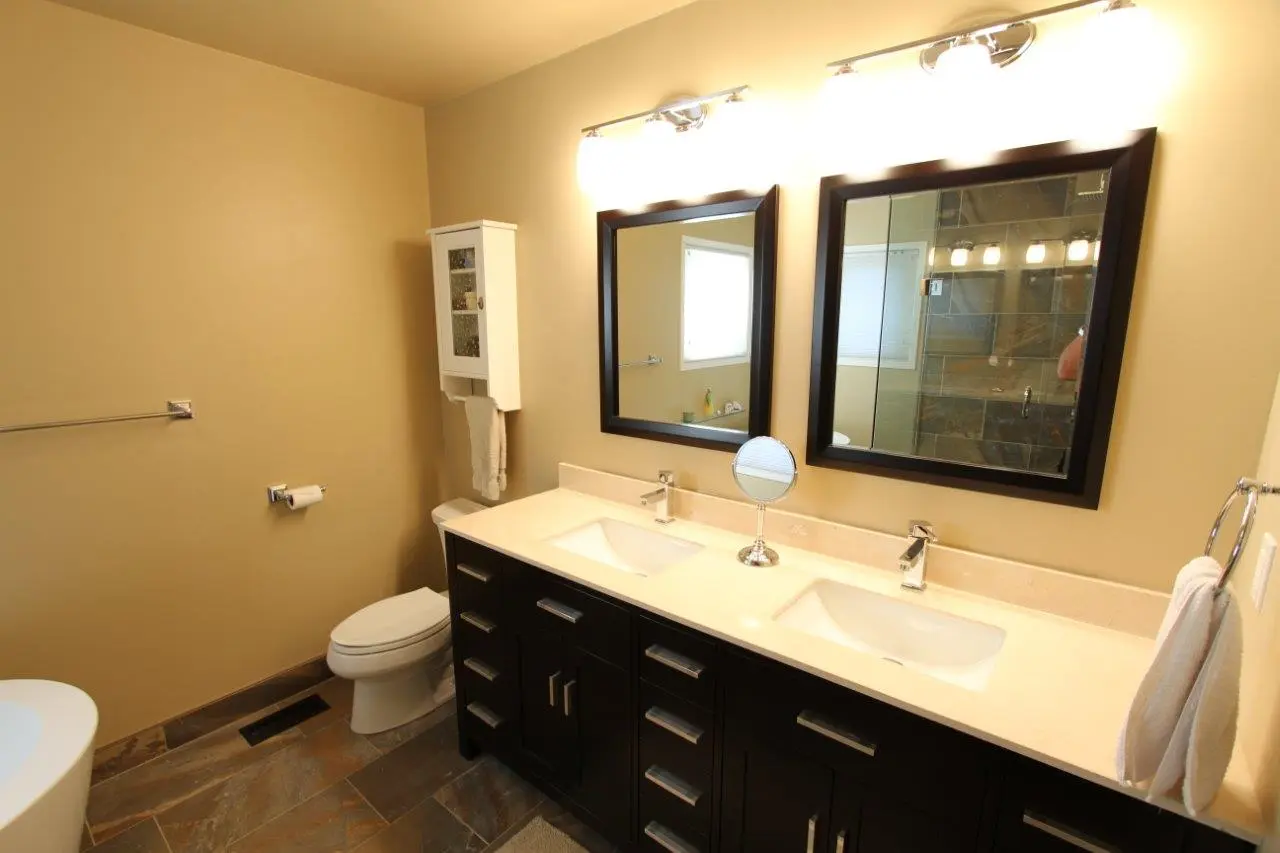 Modern bathroom with beige walls, a double sink vanity with dark cabinets and light countertop, two rectangular mirrors, a small round makeup mirror on the counter, a toilet, and a white wall-mounted cabinet with a towel hanging from it, along with towel bars on the walls.