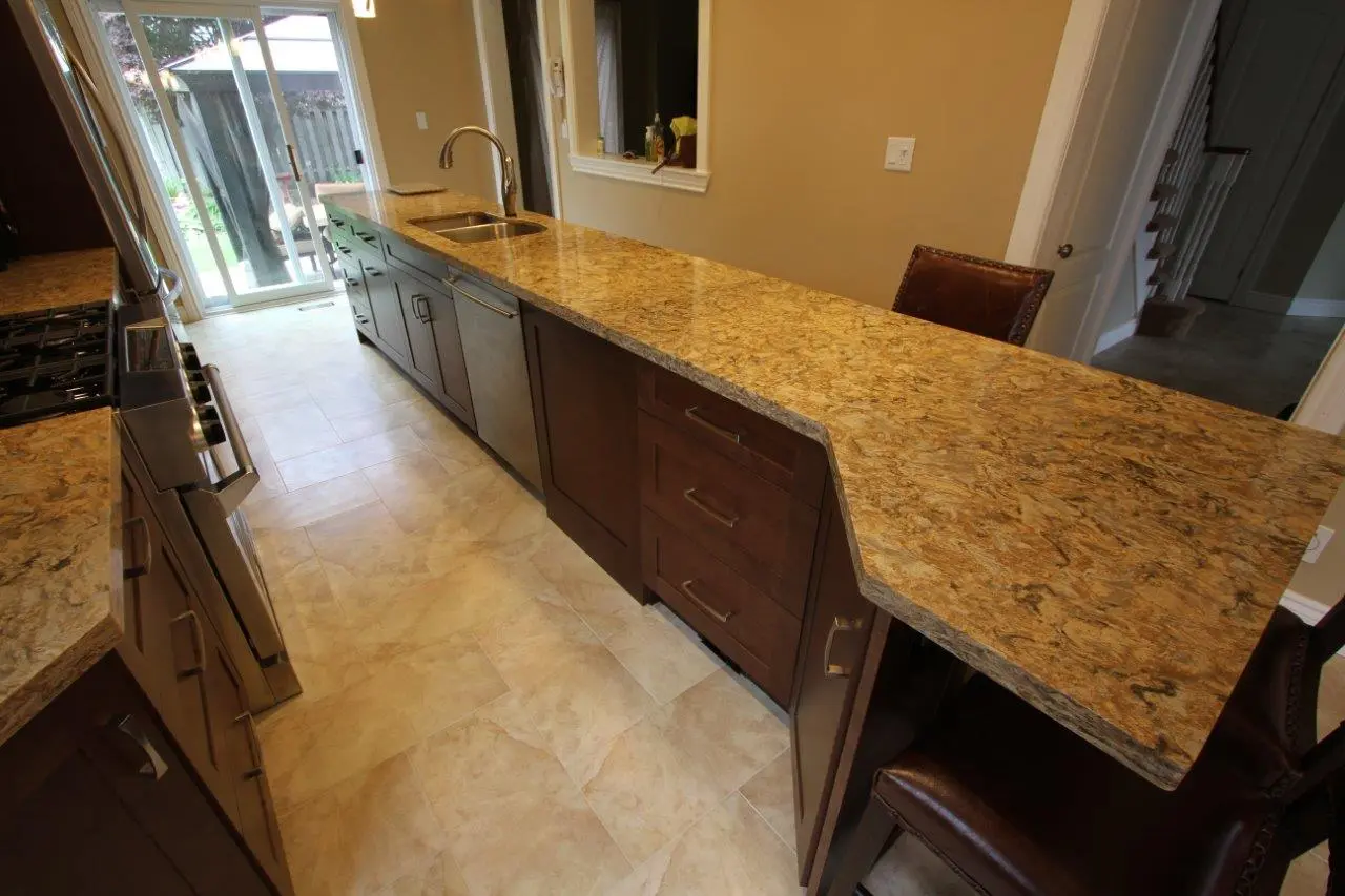 Modern kitchen with a long, polished granite countertop island featuring a built-in sink and dark wooden cabinets, tiled floor, leather bar stools, and a sliding glass door leading to an outdoor area.